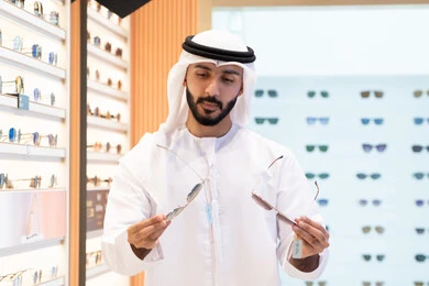 A selection of stylish sunglasses, wearing and choosing sunglasses, an Emirati Gulf Arab man wearing a kandura and ghutra choosing the right one in an Emirati store, looking at a collection of frames inside a mall