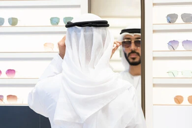 Colorful sunglasses display shelves, an Emirati Gulf Arab man wearing a kandara looking in the mirror to choose the right pair of glasses, gestures of self-confidence, offers, discounts and sales, shopping and buying from malls, the joy of marketing and shopping