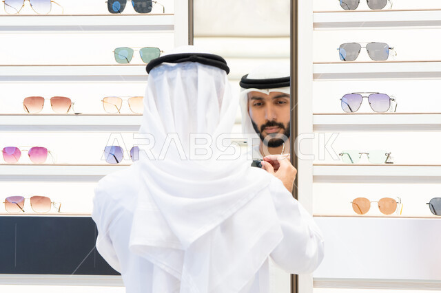 رجل عربي خليجي اماراتي يرتدي الكندره يقوم بالنظر الي المرآه لاختيار النظارة المناسبة، ايماءات الثقة بالنفس ،عروض وتخفيضات وخصومات، التسوق والشراء من المولات، متعة التسويق والتبضع ، فحص النظارات الطبيه