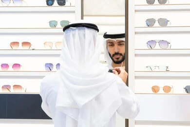 رجل عربي خليجي اماراتي يرتدي الكندره يقوم بالنظر الي المرآه لاختيار النظارة المناسبة، ايماءات الثقة بالنفس ،عروض وتخفيضات وخصومات، التسوق والشراء من المولات، متعة التسويق والتبضع ، فحص النظارات الطبيه