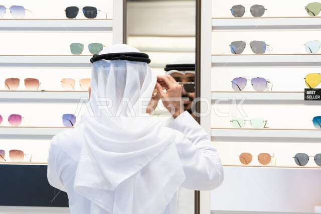 ايماءات الثقة بالنفس ،عروض وتخفيضات وخصومات، التسوق والشراء من المولات، متعة التسويق والتبضع ، فحص النظارات الطبيه ، رجل عربي خليجي اماراتي يرتدي الكندره يقوم بالنظر الي المرآه لاختيار النظارة المناسبة