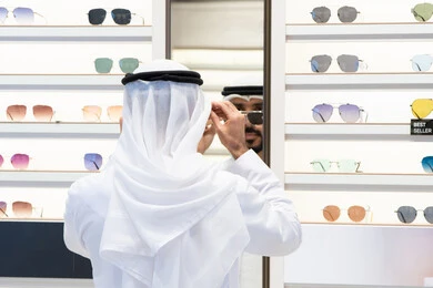 Gestures of self-confidence, offers, discounts and reductions, shopping and buying from malls, the pleasure of marketing and shopping, checking medical glasses, an Arab Gulf Emirati man wearing a kandara looking in the mirror to choose the appropriate glasses