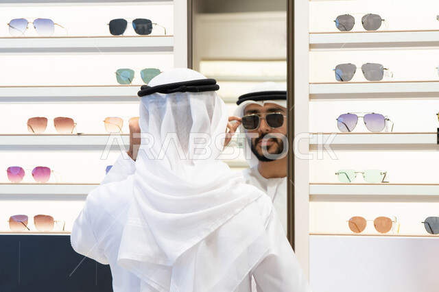 عروض وتخفيضات وخصومات، ايماءات الثقة بالنفس ، التسوق وشراء النظارات الشمسيه من المولات ، رجل عربي خليجي اماراتي يرتدي الكندره يقوم بالنظر الي المرآه لقياس النظارة المناسبة