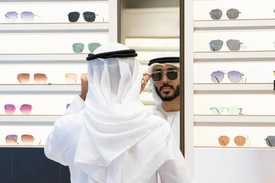 عروض وتخفيضات وخصومات، ايماءات الثقة بالنفس ، التسوق وشراء النظارات الشمسيه من المولات ، رجل عربي خليجي اماراتي يرتدي الكندره يقوم بالنظر الي المرآه لقياس النظارة المناسبة