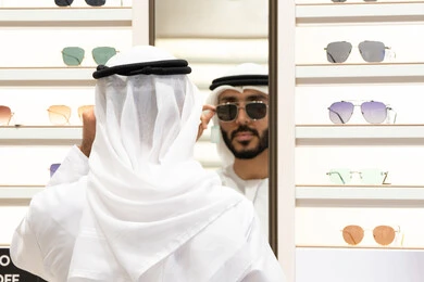 The joy of shopping, glasses display shelves inside the store, trying on black sunglasses before shopping, examining and choosing before buying, a close-up of an Emirati Gulf Arab man wearing a kandara choosing glasses