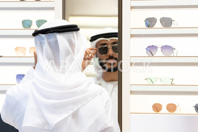 ارفف عرض النظارات داخل المحل، قياس النظارات الشمسيه السوداء قبل الشراء، متعة التسويق والشراء باختيار نظارة طبيه مناسبة، فحص النظارات الطبيه ، رجل عربي خليجي اماراتي يرتدي الكندره يقوم باختيار نظاره طبيعه