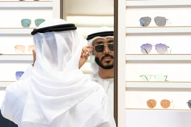 Glasses display shelves inside the store, trying on black sunglasses before buying, the pleasure of shopping and buying by choosing suitable medical glasses, examining medical glasses, an Arab Gulf Emirati man wearing a kandara choosing normal glasses