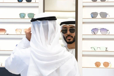 Trying on black sunglasses before buying, the joy of marketing and shopping, examining and choosing before buying, glasses display shelves inside the store, a close-up of an Arab Gulf Emirati man wearing a kandara choosing prescription glasses