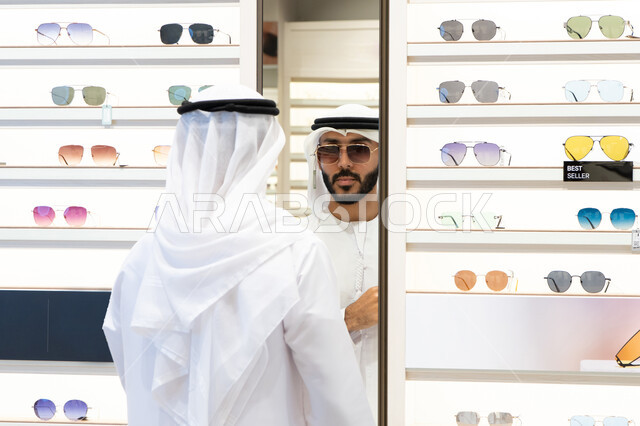 متعة التسويق والتبضع، الفحص والاختيار قبل الشراء، ارفف عرض النظارات داخل المحل، صورة مقربة لرجل عربي خليجي اماراتي يرتدي الكندره يقوم باختيار نظاره طبيه