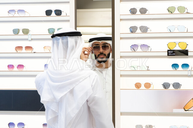 الفحص والاختيار قبل الشراء، ارفف عرض النظارات داخل المحل، متعة التسويق والشراء، صورة مقربة لرجل عربي خليجي اماراتي يرتدي الكندره يقوم باختيار نظاره طبيه