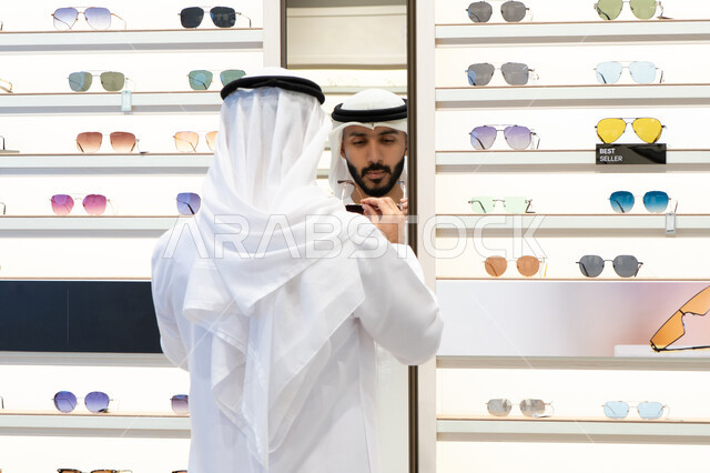 ارفف عرض النظارات داخل المحل، الفحص والاختيار قبل الشراء، متعة التسويق والشراء ، صورة مقربة لرجل عربي خليجي اماراتي يرتدي الكندره يقوم باختيار نظاره طبيه