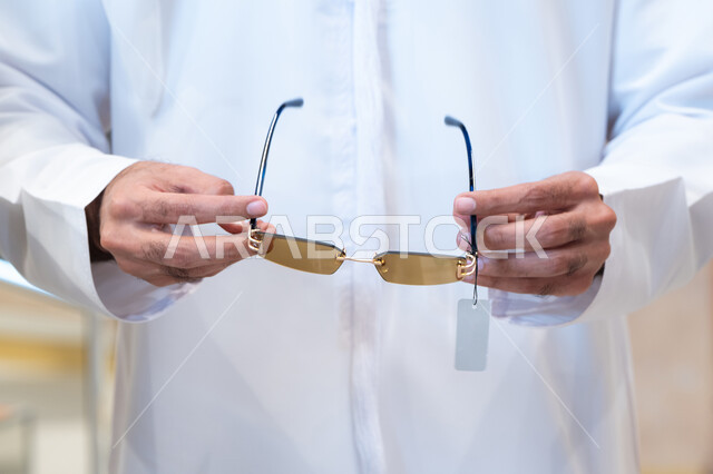 الحيرة في اختيار نظارة مناسبة ، صورة مقربة ليد رجل عربي خليجي اماراتي يرتدي الكندره يقوم باختيار نظاره طبيه، فحص النظارات قبل الشراء، متعة التسويق والشراء