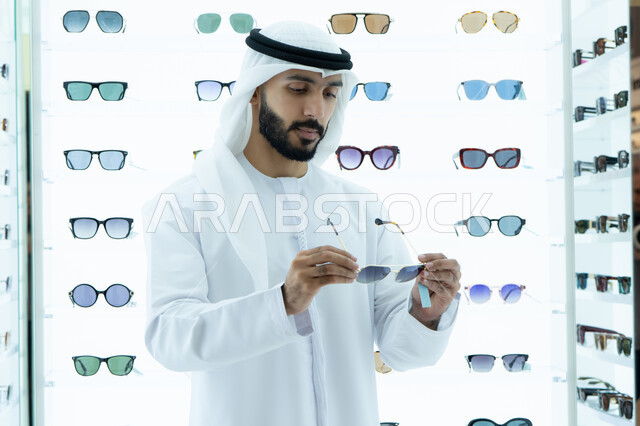 الشراء من المحلات الاماراتيه ، اختيار نظارة شمسيه مناسبة، ايماءه الثقه في النفس، رجل عربي خليجي اماراتي يرتدي الكندوره يقوم بقياس نظاره سوداء، انواع وماركات عالميه، ارفف عرض النظارات داخل المحل، متعة التسويق والشراء