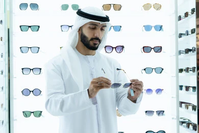 الشراء من المحلات الاماراتيه ، اختيار نظارة شمسيه مناسبة، ايماءه الثقه في النفس، رجل عربي خليجي اماراتي يرتدي الكندوره يقوم بقياس نظاره سوداء، انواع وماركات عالميه، ارفف عرض النظارات داخل المحل، متعة التسويق والشراء