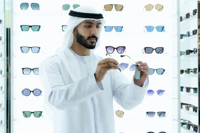 Trying on sunglasses before buying, a gesture of self-confidence, an Emirati Gulf Arab man wearing a kandura trying on black sunglasses, international brands and types, glasses display shelves inside the store, the joy of marketing and buying,