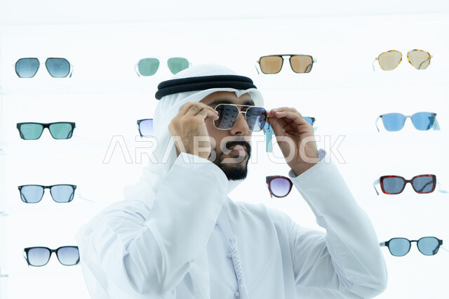 التسوق فى المولات الاماراتيه، ايماءه الثقه في النفس، رجل عربي خليجي اماراتي يرتدي الكندوره يقوم بقياس نظاره سوداء، انواع وماركات عالميه، القيام بارتداء النظارات الشمسيه قبل الشراء، ارفف عرض النظارات داخل المحل، متعة التسويق والشراء ،