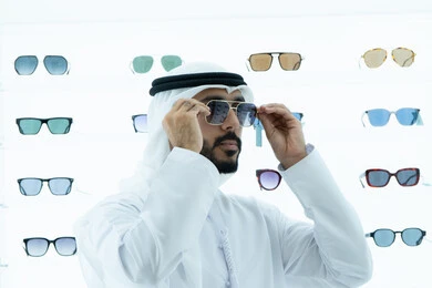 التسوق فى المولات الاماراتيه، ايماءه الثقه في النفس، رجل عربي خليجي اماراتي يرتدي الكندوره يقوم بقياس نظاره سوداء، انواع وماركات عالميه، القيام بارتداء النظارات الشمسيه قبل الشراء، ارفف عرض النظارات داخل المحل، متعة التسويق والشراء ،