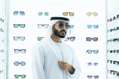رجل عربي خليجي اماراتي يرتدي الكندوره يقوم بقياس نظاره سوداء،ايماءه الثقه في النفس، انواع وماركات عالميه، القيام بارتداء النظارات الشمسيه قبل الشراء، ارفف عرض النظارات داخل المحل، متعة التسويق والشراء باختيار نظارة شمسيه مناسبة