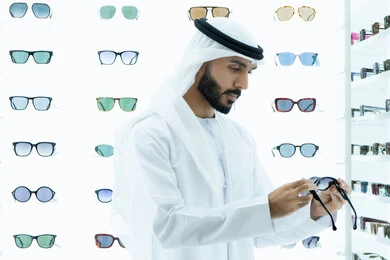 انواع وماركات عالميه، القيام بارتداء النظارات الشمسيه قبل الشراء، ارفف عرض النظارات داخل المحل، متعة التسويق والشراء باختيار نظارة شمسيه مناسبة، رجل عربي خليجي اماراتي يرتدي الكندوره يقوم بقياس نظاره سوداء،ايماءه الثقه في النفس