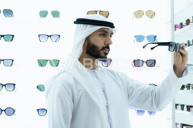 Trying on sunglasses before buying, display shelves of sunglasses inside the store, international types and brands, the pleasure of shopping and buying by choosing suitable sunglasses, an Arab Gulf Emirati man wearing a kandura and ghutra, trying on black sunglasses, a gesture of self-confidence