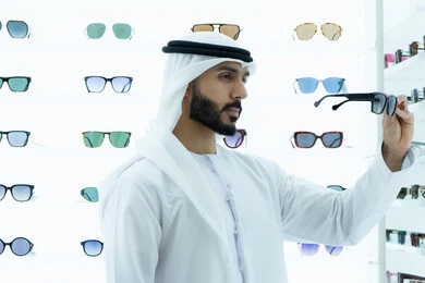Trying on sunglasses before buying, display shelves of sunglasses inside the store, international types and brands, the pleasure of shopping and buying by choosing suitable sunglasses, an Arab Gulf Emirati man wearing a kandura and ghutra, trying on black sunglasses, a gesture of self-confidence