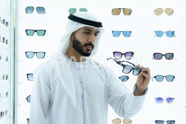 ارفف عرض النظارات داخل المحل، متعة التسويق والشراء باختيار نظارة شمسيه مناسبة، ارتداء النظارات الشمسيه قبل الشراء، رجل عربي خليجي اماراتي يرتدي الكندوره يقوم بقياس نظاره سوداء، انواع وماركات عالميه، ايماءه الثقه في النفس