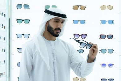 Glasses display shelves inside the store, the pleasure of shopping and buying by choosing suitable sunglasses, wearing sunglasses before buying, an Arab Gulf Emirati man wearing a kandura trying on black glasses, international types and brands, a gesture of self-confidence