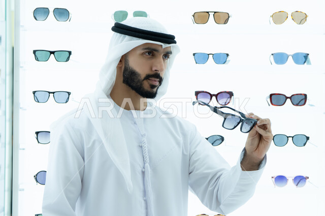 متعة التسويق والشراء باختيار نظارة شمسيه مناسبة، ارتداء النظارات الشمسيه قبل الشراء، رجل عربي خليجي اماراتي يرتدي الكندوره يقوم بقياس نظاره سوداء، ارفف عرض النظارات داخل المحل، انواع وماركات عالميه، ايماءه الثقه في النفس