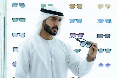 The joy of shopping and buying by choosing suitable sunglasses, wearing sunglasses before buying, an Arab Gulf Emirati man wearing a kandura trying on black sunglasses, glasses display shelves inside the store, international types and brands, a gesture of self-confidence