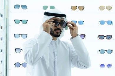 ارتداء النظارات الشمسيه قبل الشراء، متعة التسويق والشراء باختيار نظارة شمسيه مناسبة، رجل عربي خليجي اماراتي يرتدي الكندوره يقوم بقياس نظاره سوداء، ارفف عرض النظارات داخل المحل، انواع وماركات عالميه، ايماءه الثقه في النفس