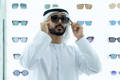 شراء النظارات الخاصة بضعف النظر، تسوق النظارات الشمسيه من المولات، شاب عربي خليجي اماراتي يرتدي الكندوره والغتره البيضاء يقوم بقياس النظارة الشمسيه المناسبة في محل نظارات، الاهتمام بالمظهر الخارجي