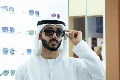 Shopping and buying sunglasses from malls, a young Emirati Gulf Arab man wearing a white kandura and ghutra trying on suitable sunglasses in an optical shop, taking care of one's outward appearance