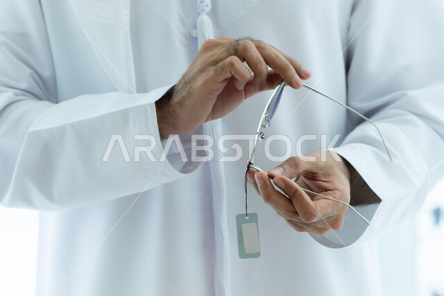 تجربة التسوق و اختيار النظارات الطبية ، الاهتمام بالمظهر الخارجي، التسوق وشراء النظارات الشمسيه من المولات ،صورة مقربه ليد شاب عربي اماراتي خليجي يرتدي الكندوره يقوم باختيار النظارة الشمسيه المناسبة في محل نظارات
