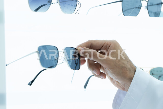 مفهوم الاناقه والاهتمام بالمظهر الخارجي ، التسوق وشراء النظارات الشمسيه من المولات ، صورة مقربه ليد شاب عربي اماراتي خليجي يرتدي الكندوره يقوم باختيار النظارة الشمسيه المناسبة في محل نظارات