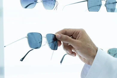 The concept of elegance and interest in external appearance, shopping and buying sunglasses from malls, a close-up photo of the hand of a young Emirati Gulf Arab man wearing a kandura choosing the appropriate sunglasses in an optical shop