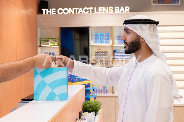 تطبيقات التسوق والشراء الالكتروني عبر الهاتف، تسوق وشراء أحدث منتجات النظارات العصرية الحديثة, رجل عربي خليجي اماراتي يحمل كيس ورقي ملون، اقتناء وشراء هدايا من احدي المولات الاماراتيه، إتمام عملية الدفع باستخدام الهاتف المحمول