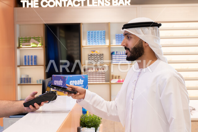 An Emirati Gulf Arab man makes an electronic payment, purchasing gifts from an Emirati mall, using online shopping and purchasing applications via phone, completing the payment process using a mobile phone.