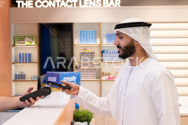 اقتناء وشراء هدايا من احدي المولات الاماراتيه، تطبيقات التسوق والشراء الالكتروني عبر الهاتف، إتمام عملية الدفع باستخدام الهاتف المحمول، رجل عربي خليجي اماراتي يقوم بالدفع الالكتروني