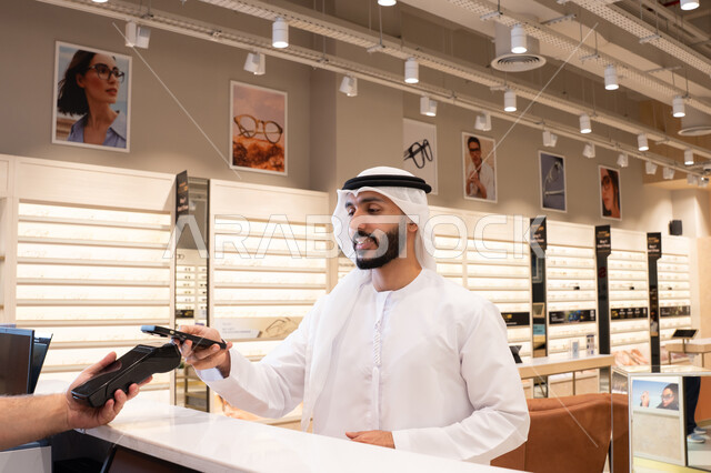 إتمام عملية الدفع باستخدام الهاتف المحمول، تطبيقات التسوق والشراء الالكتروني عبر الهاتف، اقتناء وشراء هدايا من احدي المولات الاماراتيه، رجل عربي خليجي اماراتي يقوم بالدفع الالكتروني