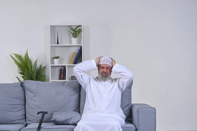 Stress and psychological pressure, portrait of an elderly Arab Gulf Emirati man wearing a kandura and ghutra sitting on a sofa, placing his hands on his head and closing his eyes with gestures of severe pain, exhaustion and loss of concentration, feeling a headache and head pain, feeling of migraine and migraine, white background