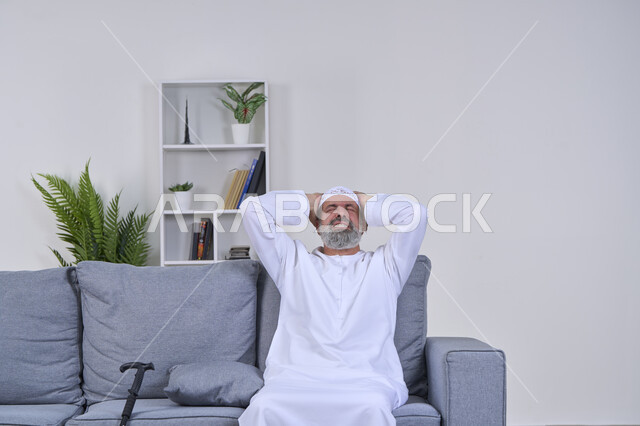 الاحساس بالشقيقية والصداع النصفي, الاجهاد والضغط النفسي, بورتريه لرجل مسن عربي خليجي اماراتي يرتدي الكندورة والغترة يجلس على كنبه ويضع يديه على رأسه ويغمض عينيه بإيماءات الألم الشديد, الإرهاق وفقدان التركيز، الشعور بالصداع وألم الرأس, خلفية بيضاء