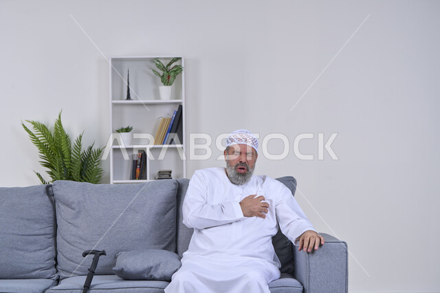 Fatigue, exhaustion and psychological pressure, feeling of increased heart rate, feeling of angina, portrait of an elderly Arab Gulf Emirati man wearing a kandura and hat sitting on a sofa with his hand on his heart and feeling a bad state of tingling and pain, heart disorders and diseases, white background