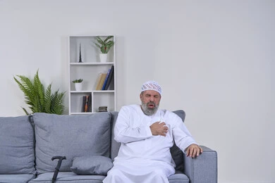 Fatigue, exhaustion and psychological pressure, feeling of increased heart rate, feeling of angina, portrait of an elderly Arab Gulf Emirati man wearing a kandura and hat sitting on a sofa with his hand on his heart and feeling a bad state of tingling and pain, heart disorders and diseases, white background