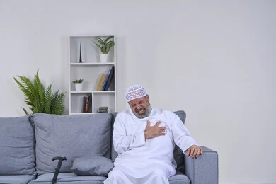 الشعور في زيادة معدل ضربات القلب, التعب والإرهاق والضغوطات النفسية, الاحساس بذبحه صدريه، بورتريه لرجل مسن عربي خليجي اماراتي يرتدي الكندورة والقبعة يجلس على كنبة ويضع يده على قلبه ويشعر بحالة سيئة من الوخز والألم, اضطرابات وأمراض قلبية, خلفية بيضاء