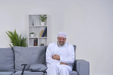 آلام المعدة والأمعاء، إيماءات الوجه تدل على الاضطراب والتألم، ضرورة الذهاب للطبيب والحصول على العلاج، بورتريه لرجل كبير السن عربي خليجي إماراتي يرتدي الكندورة والقبعة يجلس على كنبه ويضع يديع على بطنه بإيماءات الألم الشديد, خلفية بيضاء