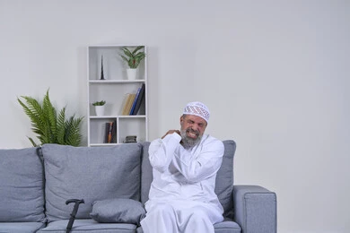 Facial expressions indicating distress and pain, the need to go to the doctor and get treatment, portrait of an elderly Arab Gulf Emirati man wearing a kandura and a hat sitting on his sofa and putting his hands on his shoulder with gestures of severe pain, back, bone and spine pain, white background