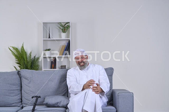 Joint, bone and nerve pain, facial expressions indicating distress and pain, the need to go to the doctor and get treatment, portrait of an elderly Arab Gulf Emirati man wearing a kandura and a hat sitting on his sofa and holding his knee with gestures of severe pain, white background