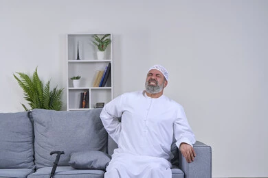 The necessity of going to the doctor and getting treatment, a portrait of an elderly Arab Gulf Emirati man wearing a ghutra and a hat sitting on his sofa and putting his hand on his back with gestures of severe pain, back, bone and spine pain, facial gestures indicating distress and pain, white background
