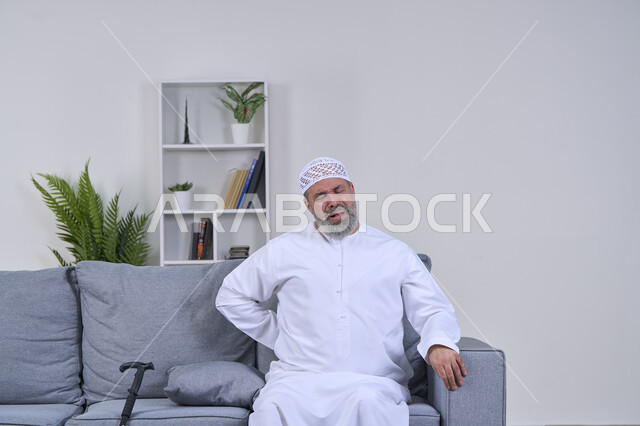 ضرورة الذهاب للطبيب والحصول على العلاج، بورتريه لرجل كبير السن عربي خليجي إماراتي يرتدي الغترة والقبعة يجلس على كنبه ويضع يده علىى ظهره بإيماءات الألم الشديد, آلام الظهر والعظام والعمود الفقري، إيماءات الوجه تدل على الاضطراب والتألم، خلفية بيضاء