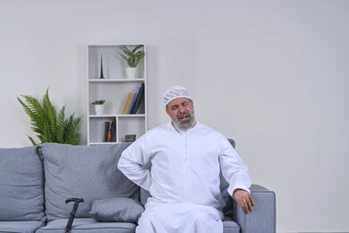 The necessity of going to the doctor and getting treatment, a portrait of an elderly Arab Gulf Emirati man wearing a ghutra and a hat sitting on his sofa and putting his hand on his back with gestures of severe pain, back, bone and spine pain, facial gestures indicating distress and pain, white background